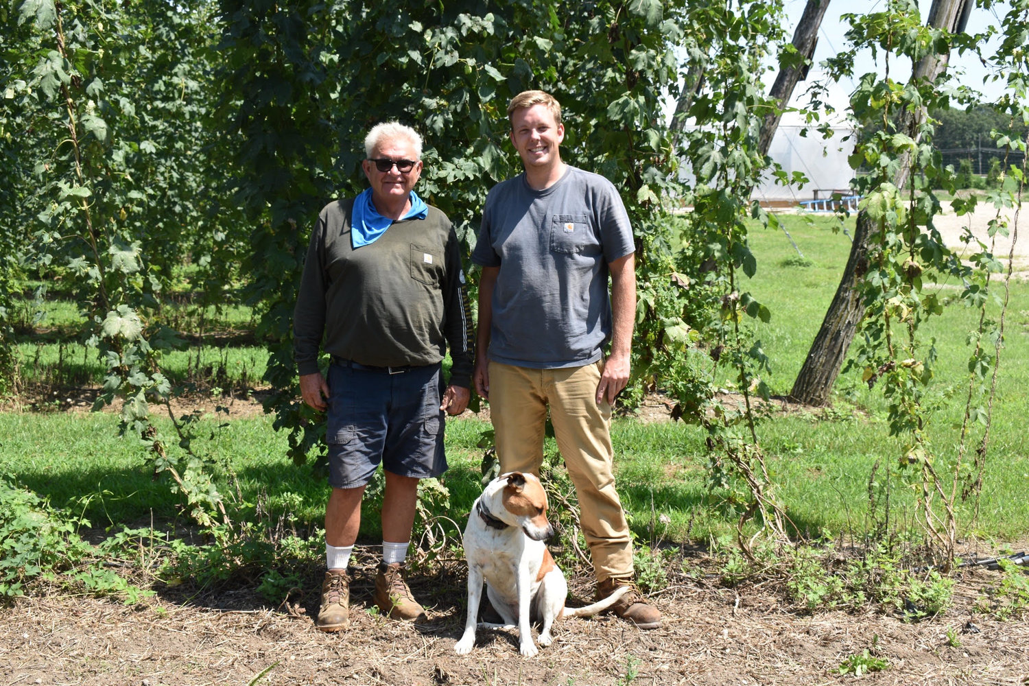 Mark Carroll (left) is pictured with Ryan Andoos (right) and Shelby (front, center) in front of a hops plant.
